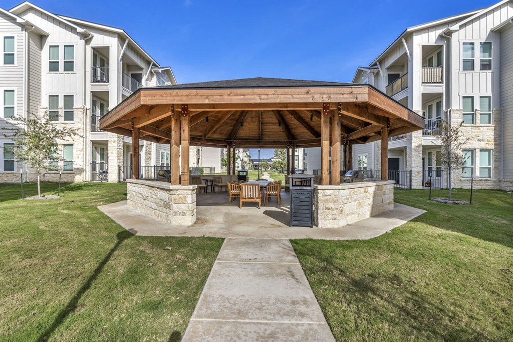 A gazebo-like structure sits in the middle of a grassy area in front of apartment buildings.