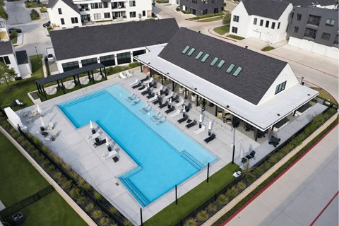 A large swimming pool with lounge chairs and umbrellas is surrounded by a white building and other buildings in the background.