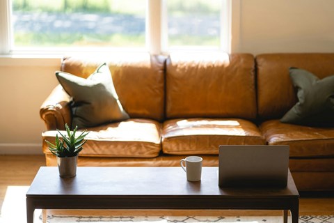 A brown leather couch with a laptop and a cup on a wooden table in front of it.