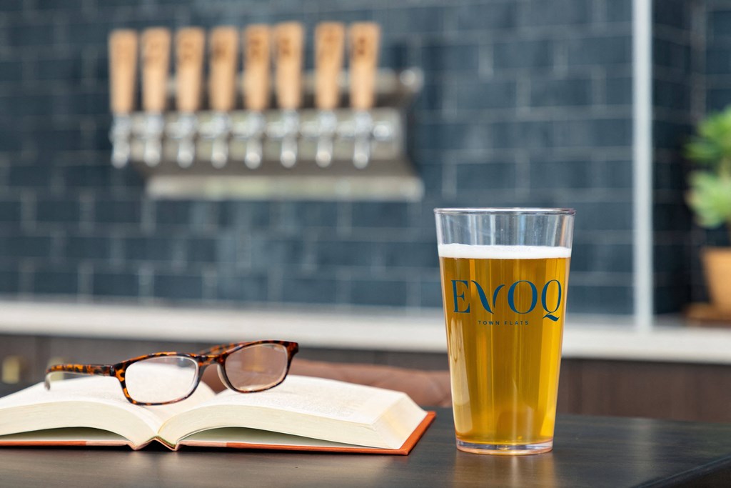 A glass of beer with the word EVOQ on it sits on a table next to an open book and a pair of glasses.
