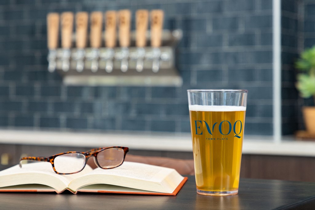 A glass of beer with the word EVOQ on it sits on a table next to an open book and a pair of glasses.