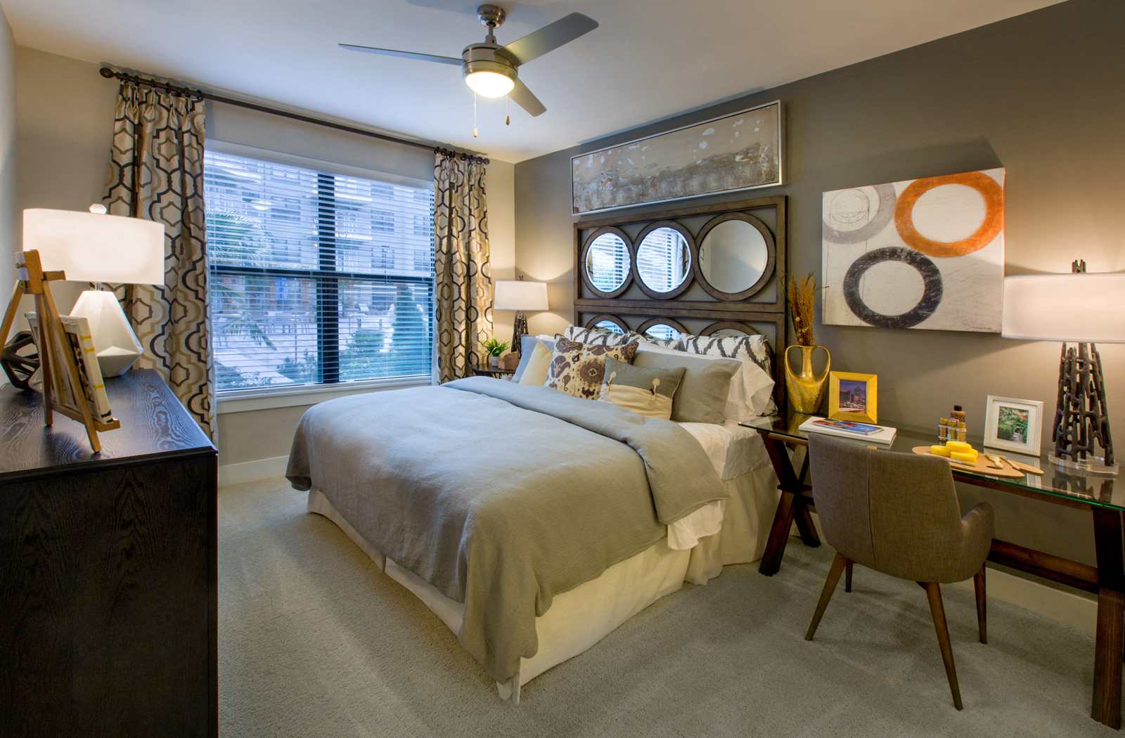 Carpeted bedroom with a bed, nightstand, desk and chair, dresser, and large window facing the pool area.