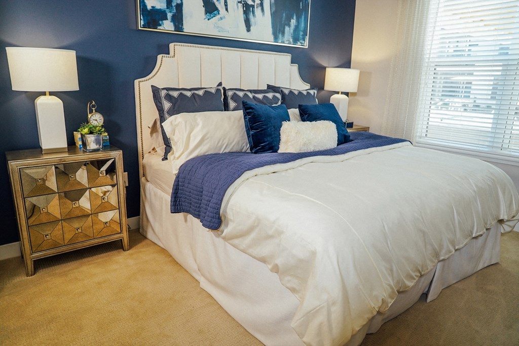 Bed between 2 nightstands near a large window in a carpeted bedroom. 