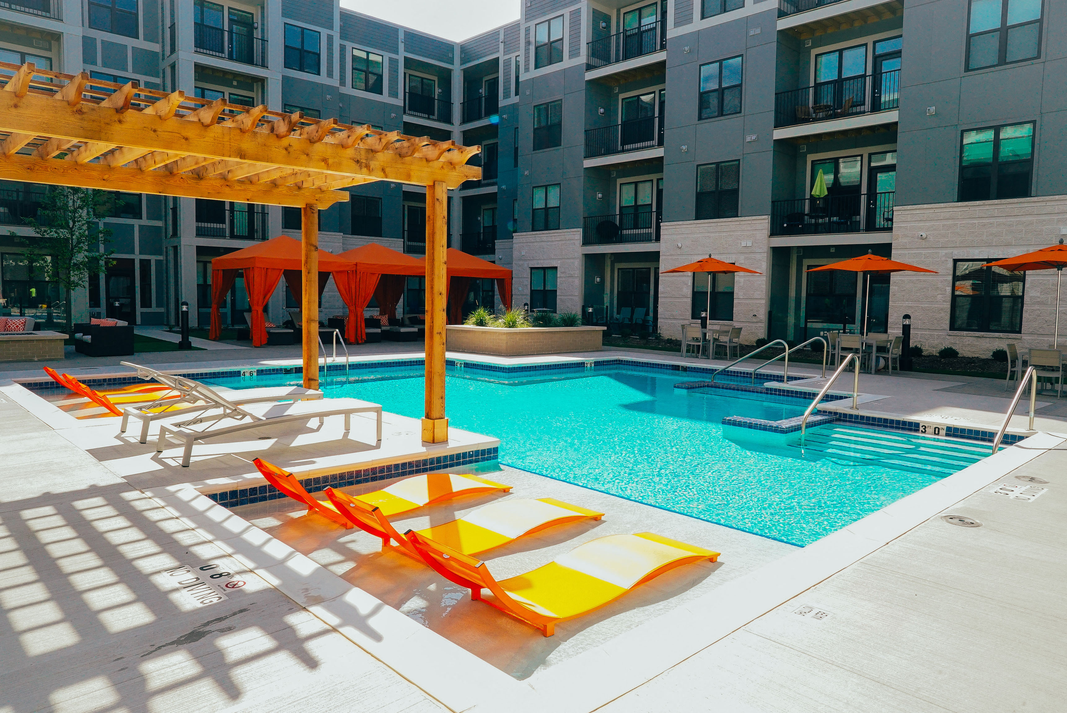 Outdoor pool area with multiple lounge chairs and shaded seating areas, surrounded by apartments. 