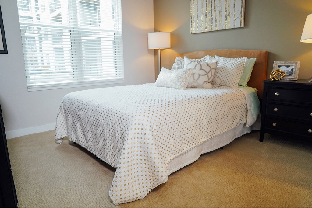 Carpeted bedroom with a bed in the middle, next to a nightstand, a standing lamp, and a large window. 