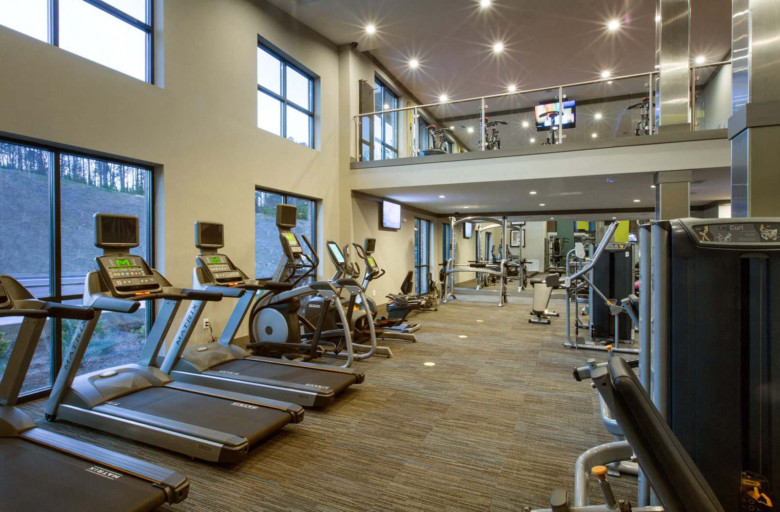 Two-level fitness center with various workout equipment and large windows facing the outdoors. 