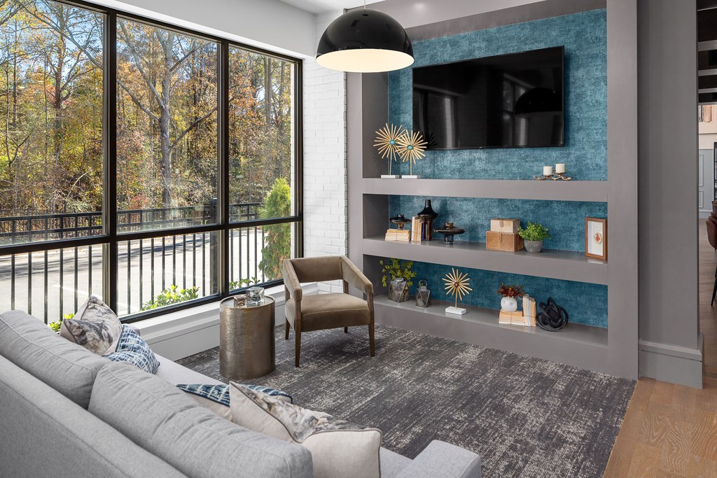 A modern living room with a grey sofa and a large window overlooking a tree-lined street.