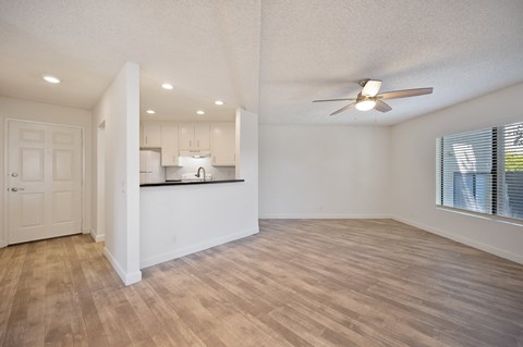 an empty living room and kitchen with a ceiling fan, vinyl wood flooring, white walls, and a breakfast bar.