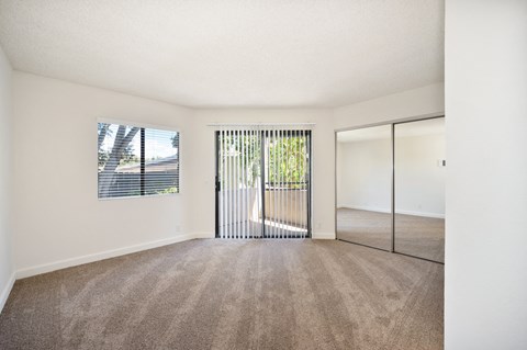 A bedroom with carpet, a window, a sliding glass door to a balcony, and a mirrored sliding closet door.