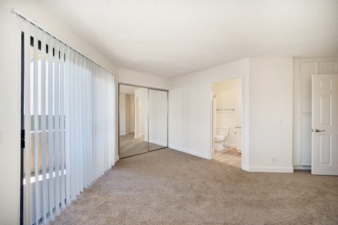 an empty bedroom with a sliding glass door to a balcony, carpet, white walls, and mirrored sliding closet doors.