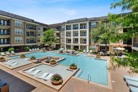 a fire pit surrounded by chairs and a pool in front of an apartment building. there are pool lounge chairs and plants in the foreground