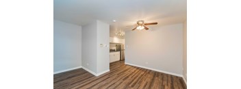 A room with a ceiling fan and wooden flooring with a kitchen in the background.