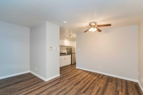 A room with a ceiling fan and wooden flooring with a kitchen in the background.