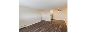 A room with wooden style flooring and a ceiling fan with a peak view of a kitchen in the background.