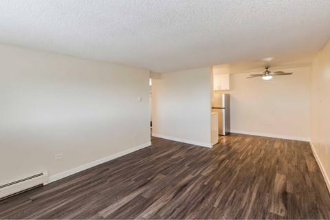 A room with wooden style flooring and a ceiling fan with a peak view of a kitchen in the background.
