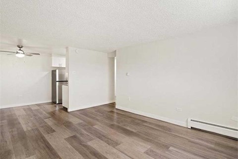 A room with a wooden floor and a ceiling fan and a kitchen with a fridge in the background.