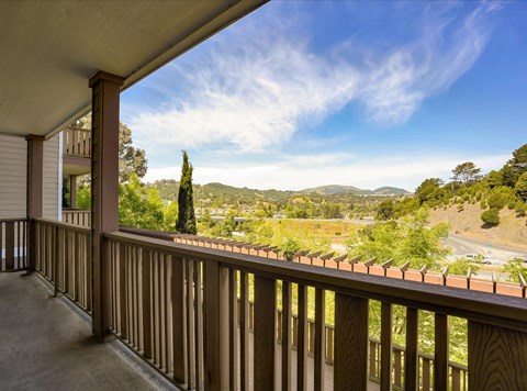 View from a balcony overlooking a scenic landscape. Lush green hills and trees are visible under a blue sky with wispy clouds. The foreground features a wooden railing, and the overall atmosphere conveys a peaceful, natural setting.