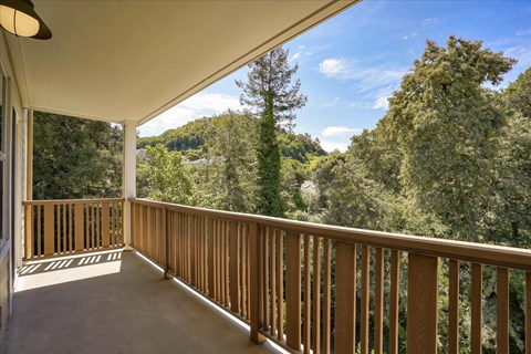 A spacious balcony with a wooden railing overlooking a lush green landscape, featuring tall trees and a clear blue sky with scattered clouds. The scene conveys a peaceful outdoor space ideal for relaxation and enjoying nature.