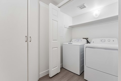 Apartment interior full size washer and dryer in white. Laundry area has bi-fold doors and ample shelving above the washer and dryer.