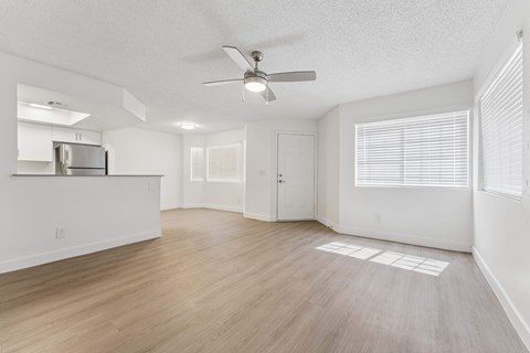 Apartment interior living room area with light brown vinyl flooring, ceiling fan, white walls, and slight view of kitchen counter and dining room area.