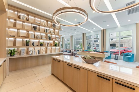Modern lounge area featuring a light wood bookshelf filled with decorative items and plants, stylish pendant lighting, a spacious countertop, and a seating area with colorful chairs. Large windows let in natural light, while a view of parked cars is visible outside.
