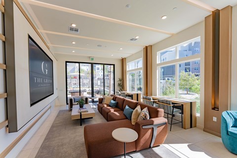 Interior of a modern lounge featuring a brown sectional sofa, a light-colored coffee table, and large windows letting in natural light. A large screen displays "THE GRAND." There are additional seating options, a workspace with desks, and greenery visible outside. The design is contemporary and inviting.