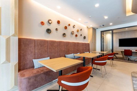 Cozy interior of a modern café featuring a brown upholstered bench, wooden tables, and orange chairs. The wall is decorated with circular wall art in various colors, and there is a glass partition leading to a room with a television. The lighting is warm and inviting, creating a comfortable atmosphere.
