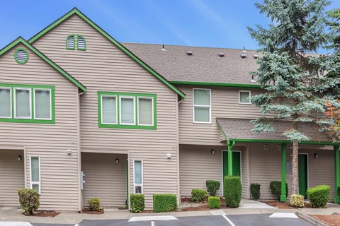 Community exterior with tan siding and green and white accents with shrubs and pine trees.