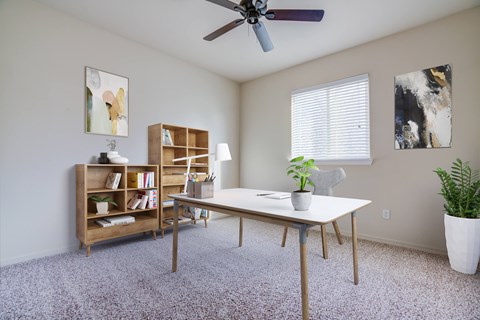A bedroom with carpet, a ceiling fan, and a window with blinds. The room is staged as an office with shelving, a desk, and a chair.