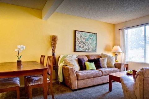 A living room carpet, a yellow accent wall, and a window with blinds. The room is staged with a couch, coffee table, a painting, and a dining room table set for 4.