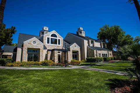 Exterior community clubhouse entry with stone facade, shrubs, and lawn, wide view.