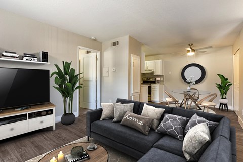 A living room with wood style plank flooring, beige walls, and a view of the dining room with a ceiling fan. The room is staged with a tv, a couch, a rug, and a plant.