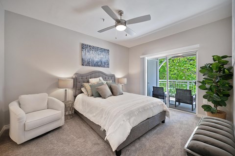 A bedroom with carpet, a ceiling fan, and a sliding glass door to the patio he room is staged with a bed, a chair, and nightstands with lamps on them.