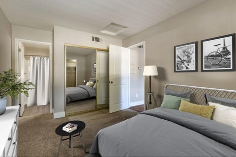 A bedroom with a carpet, beige walls, and a mirrored closet door. The room is staged with a bed, a lamp, picture frames above the bed, and a dresser.
