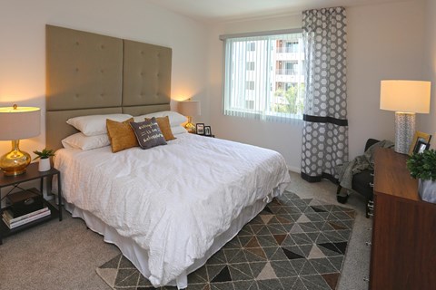 A bedroom with light colored carpet, white walls, and a window. The room is staged with a bed, nightstands, and a dresser.