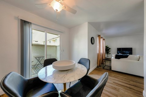 A dining room with white walls, wood style plank flooring, a ceiling fan, and a sliding glass door to a balcony. The area is staged with a dining room table set for 4.