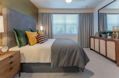 A cozy bedroom featuring a bed with gray and colorful decorative pillows, a neutral-colored blanket, and a stylish headboard. There's a wooden nightstand with a clock, sheer curtains covering a window, and a large mirror above a wooden dresser. The decor is modern with earthy tones and inviting textures.