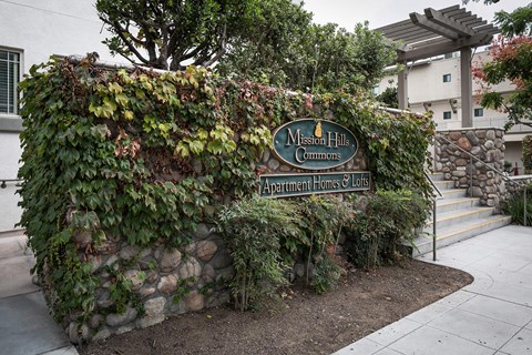A sign for Mission Hills Commons Apartment Homes & Lofts is surrounded by greenery.