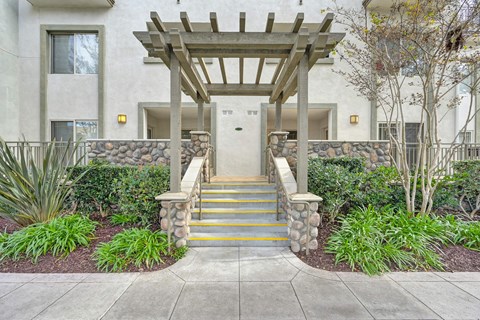 A white building with a wooden pergola and a stone wall with steps leading up to the entrance.