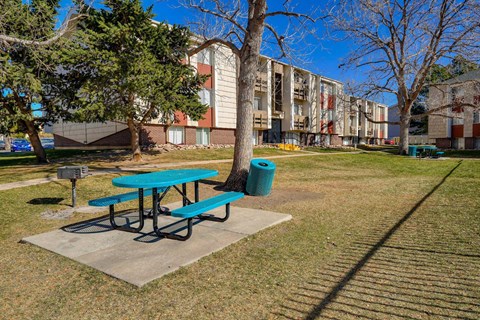 a picnic area with a blue picnic table with black legs, a barbecue, a blue trash can, in front of an apartment building and trees.
