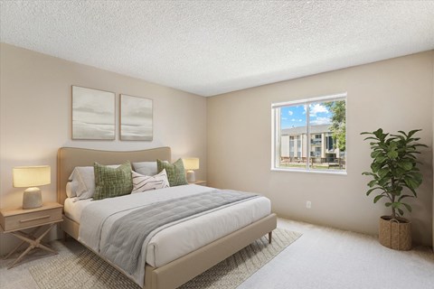 A bedroom with a large bed, a nightstand with a lamp, a window with a view of a building and trees outside, and a potted plant.