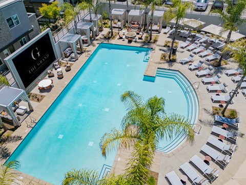 Aerial view of a luxurious outdoor swimming pool surrounded by palm trees, lounge chairs, and shaded cabanas. The pool features a unique shape with a light blue water surface. In the background, a large sign reads "The Grand." The atmosphere is bright and inviting, ideal for relaxation.