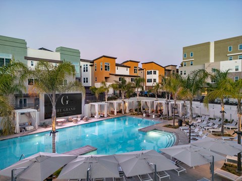 A modern swimming pool surrounded by lounge chairs with tropical palm trees. The pool area features sleek, contemporary architecture with colorful apartment buildings in the background. Soft lighting creates a relaxing atmosphere, making it an inviting space for leisure and relaxation.