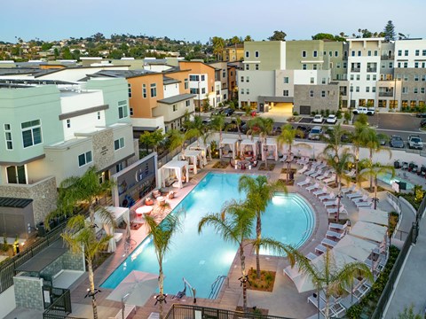 Aerial view of a modern apartment complex featuring a large swimming pool surrounded by palm trees and lounge chairs. The area is well lit, showcasing the pool and surrounding buildings. The setting reflects a vibrant, upscale community atmosphere.