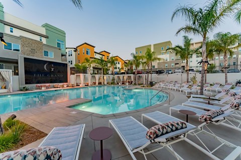 A modern swimming pool surrounded by lounge chairs with tropical palm trees. The pool area features sleek, contemporary architecture with colorful apartment buildings in the background. Soft lighting creates a relaxing atmosphere, making it an inviting space for leisure and relaxation.
