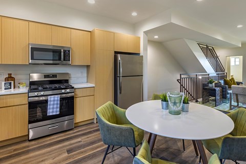 A modern kitchen with stainless steel appliances, wooden cabinets, quartz countertops, and vinyl flooring. The room is staged with a dining room table and chairs.