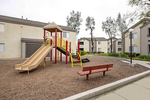 A playground with a yellow and red slide, a wooden swing set, and a red bench.