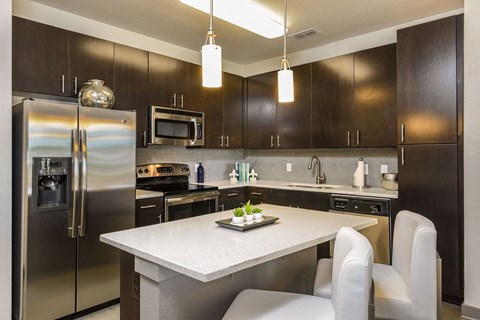 A modern kitchen featuring dark wood cabinets, stainless steel appliances including a refrigerator and oven, gray countertops, and a small dining area with white chairs and a decorative centerpiece. Bright overhead lighting enhances the contemporary design.