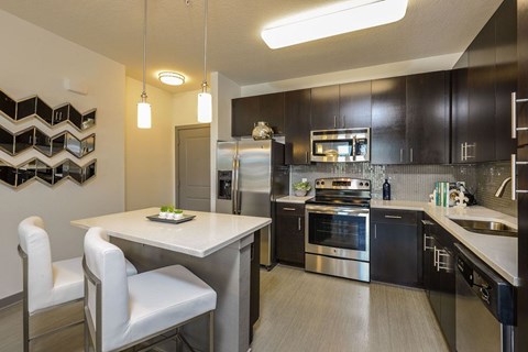 Modern kitchen featuring dark wood cabinetry, stainless steel appliances, and a stylish backsplash. A small dining area with two white chairs and a table is in the foreground. Decorative wall art can be seen on the left, adding to the contemporary aesthetic. Bright lighting enhances the warm atmosphere.