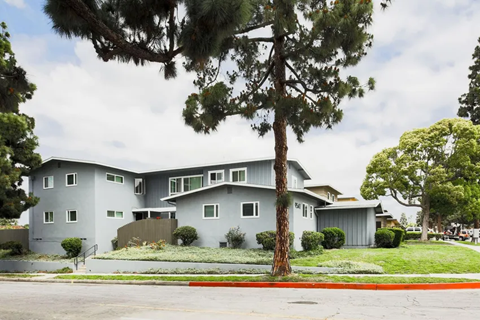 A modern, multi-story gray apartment building surrounded by greenery. The structure features large windows and a landscaped yard with shrubs. Tall trees provide shade in the vicinity, and a gray sidewalk runs along the front. The sky is partly cloudy, creating a serene atmosphere.